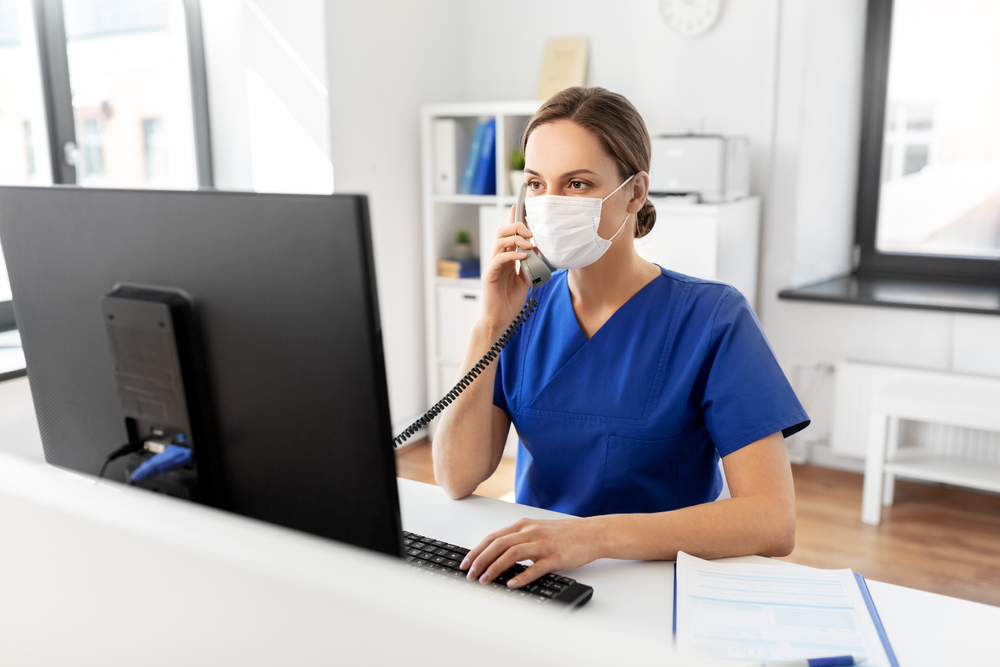Masked medical assistant talking on the phone in front of a computer